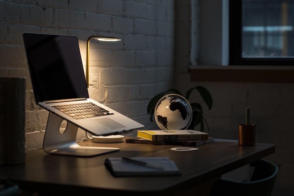 A laptop raised on an aluminium stand beside a desk lamp